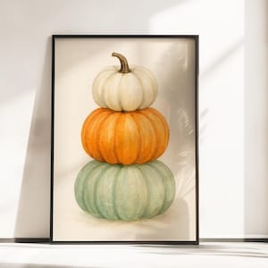 May include: A framed watercolor print featuring three stacked pumpkins. The top pumpkin is white, the middle one is orange, and the bottom one is a muted green. The pumpkins have a realistic, textured appearance, and the artwork is set against a neutral background.