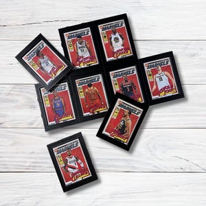 May include: A collection of framed Marvels basketball trading cards featuring various players. Each card has a red background with player images and text. The frames are black, and the cards are arranged on a white wooden surface.