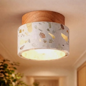 May include: A flush-mount ceiling light fixture with a wooden top and a cylindrical body. The body is made of speckled concrete with yellow, brown, and gray accents. The light is on, emitting a warm glow.