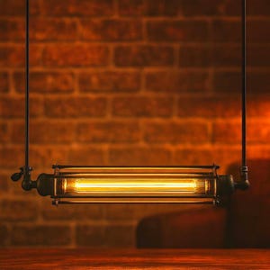 Puede incluir: Lámpara colgante de estilo vintage con una bombilla larga y rectangular dentro de un marco metálico. Se suspende de varillas negras y emite una luz amarilla cálida. El fondo es una pared de ladrillos.