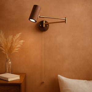 Könnte beinhalten: Eine Wand-Leselampe mit einem Lampenschirm aus dunklem Holz und einem Arm und Akzenten in Messingfarbe. Die Lampe ist auf einer kreisförmigen Basis aus dunklem Holz montiert. Eine Zugkette hängt von der Basis herab. Die Lampe befindet sich vor einer warmen, braunen Wand.