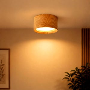 Puede incluir: Una lámpara de techo cilíndrica con un acabado de veta de madera natural de color marrón claro. El accesorio está montado al ras del techo y emite un brillo cálido y brillante hacia abajo, iluminando el espacio directamente bajo la luz.