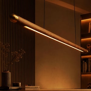 May include: A linear pendant light fixture featuring a long, cylindrical wooden body with a smooth finish. The fixture is capped with a metallic gold end piece and contains a continuous, warm-toned LED light strip running along its underside.