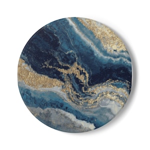 May include: Round coaster with an abstract design featuring swirling shades of blue, navy, and white, accented with gold metallic streaks. The coaster has a glossy finish and a clean, modern aesthetic.