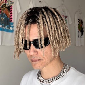以下が含まれることがあります： 黒いサングラスとシルバーのチェーンネックレスを身に着けた人。ブロンドのドレッドヘアで白いTシャツを着ています。背景の壁には白いTシャツがいくつか掛かっています。