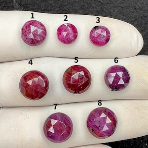 Puede incluir: Una colección de ocho gemas redondas facetadas en varios tonos de rojo y rosa. Las gemas se muestran sobre una superficie blanca. Cada piedra tiene un patrón único de facetas, creando un efecto brillante. Las gemas están numeradas del 1 al 8.