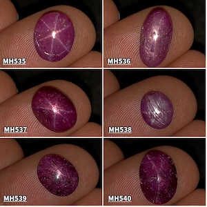Könnte beinhalten: Sechs ovale, polierte Rubin-Edelsteine. Die Edelsteine haben eine tiefrote bis violette Farbe, wobei einige einen Sterneneffekt aufweisen. Jeder Stein ist mit einem weißen Textcode beschriftet: MH535, MH536, MH537, MH538, MH539 und MH540.
