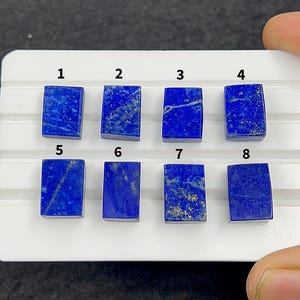 May include: Eight rectangular lapis lazuli gemstones arranged in two rows on a white display tray. Each stone features a deep blue color with varying patterns of gold pyrite flecks and white calcite veins. The stones are numbered one through eight.