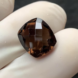 Pode incluir: Uma gema facetada, com corte almofada, de cor castanho fumado intenso. A gema é segurada por uma mão enluvada, mostrando sua clareza e múltiplas superfícies refletoras. A forma da gema é quadrada com cantos arredondados.