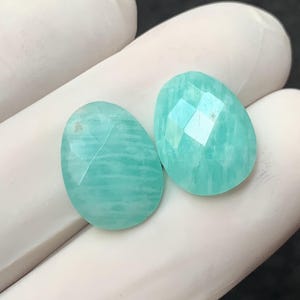 Peut inclure: Deux pierres précieuses taillées à facettes en forme de goutte d'eau. Les pierres sont de couleur turquoise clair avec de subtiles variations de tons et des motifs internes visibles. Les facettes créent un effet scintillant. Les pierres sont posées sur une surface blanche.