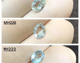 May include: Three aquamarine gemstones are displayed. The top two are pear-shaped, and the bottom one is oval. Each gem is a light blue color and appears to be faceted. The text "MH219", "MH220", and "MH223" are printed above each gem.