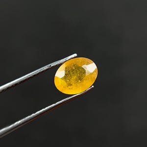 Puede incluir: Una piedra preciosa facetada de forma ovalada con un tono amarillo anaranjado vibrante. La piedra presenta una superficie pulida con inclusiones internas y está sujeta por unas pinzas de metal plateado, mostrando su calidad translúcida.