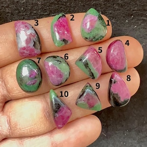 May include: A collection of ten polished ruby zoisite gemstones in various shapes. The stones display a mix of pink, green, and black colors. Each stone is unique, with varying patterns and sizes, suitable for jewelry making.
