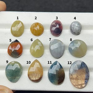 Può includere: Una collezione di dodici gemme sfaccettate in varie forme e colori, tra cui giallo, arancione, rosso, grigio e blu. Le gemme sono disposte in una griglia, mostrando i loro tagli e tonalità unici. Ideale per la creazione di gioielli.