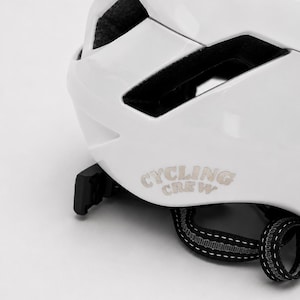 Könnte beinhalten: Ein glänzender weißer Fahrradhelm mit dem Schriftzug "CYCLING CREW" in einer hellen Schriftart an der Seite. Der Helm verfügt über schwarze Belüftungsschlitze, ein verstellbares schwarzes Gurtsystem und eine sichtbare schwarze Kunststoffschnalle.