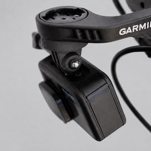 Może przedstawiać: Czarna kamera sportowa zamontowana na czarnym uchwycie rowerowym marki Garmin. Kamera jest ustawiona pod kątem, ukazując swój prostokątny korpus, okrągłą osłonę obiektywu oraz nazwę marki Garmin nadrukowaną na biało na ramieniu montażowym.