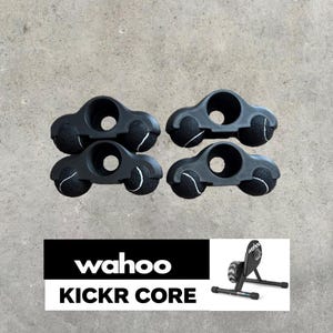 Könnte beinhalten: Vier schwarze Kunststoffhalterungen, jede mit zwei integrierten schwarzen Tennisbällen mit weißen Nähten. Diese Halterungen sind für den Wahoo KICKR CORE Indoor-Fahrradtrainer konzipiert, der in einer kleinen Grafik mit dem Text Wahoo KICKR CORE zu sehen ist.