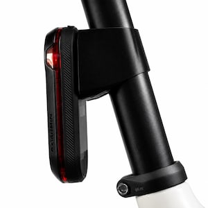 May include: A black Garmin bicycle tail light mounted to a vertical black seat post. The device features a slim, rectangular profile with a red lens strip along the side and a secure plastic mounting bracket attached to the cylindrical post.