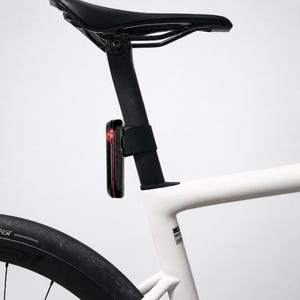 Puede incluir: Un cuadro de bicicleta blanco con una tija de sillín y un sillín negros. Una pequeña luz trasera rectangular negra para bicicleta, con un indicador rojo brillante, está montada en la tija, justo debajo del sillín en el cuadro.