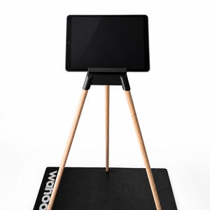 Op de afbeelding: Een zwarte tablet gemonteerd op een statief met drie lichtgekleurde houten poten en een zwarte kunststof bovenbeugel. De standaard staat op een zwarte rechthoekige mat met de witte tekst wahoo langs de linkerzijde gedrukt.
