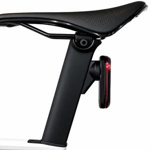May include: A black bicycle saddle mounted on a black seat post with a small rectangular red LED tail light attached to the rear. The saddle features a perforated texture on the rear section and is connected to a white bicycle frame base.