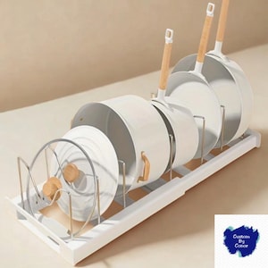 Può includere: Un organizer da cucina bianco in metallo e legno che contiene diverse pentole, padelle e coperchi bianchi. Le pentole e le padelle hanno manici in legno. L'organizer è bianco e ha divisori in metallo.