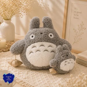 May include: Two gray and white plush toys shaped like creatures with rounded ears, large white eyes, and a white belly featuring gray chevron markings. Each plush toy holds a small, beige, star-shaped fabric accessory by a thin string.