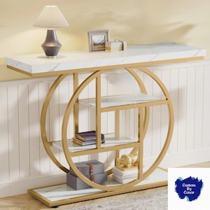 Peut inclure: Une console moderne avec un plateau en marbre blanc et une structure en m&eacute;tal dor&eacute;. La table pr&eacute;sente un design d'&eacute;tag&egrave;re circulaire avec plusieurs &eacute;tag&egrave;res en verre. Une lampe, un cadre photo et des objets d&eacute;coratifs sont expos&eacute;s.