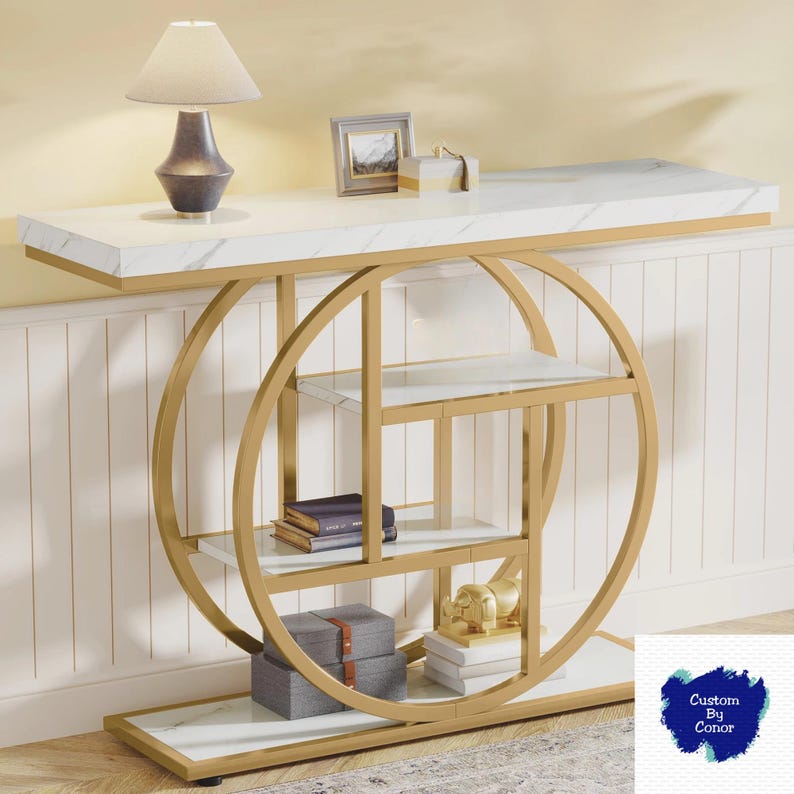 Peut inclure: Une console moderne avec un plateau en marbre blanc et une structure en m&eacute;tal dor&eacute;. La table pr&eacute;sente un design d'&eacute;tag&egrave;re circulaire avec plusieurs &eacute;tag&egrave;res en verre. Une lampe, un cadre photo et des objets d&eacute;coratifs sont expos&eacute;s.