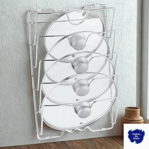 Puede incluir: Un estante de metal plateado para tapas de ollas que sostiene cuatro tapas de vidrio con perillas plateadas. El estante está montado en una pared gris claro. Las tapas son transparentes y redondas.