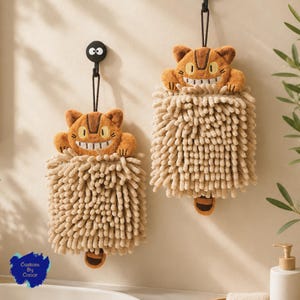 May include: Two tan chenille hand towels featuring an embroidered cat character with a wide grin, yellow eyes, and a striped head. Each towel has a hanging loop and a small brown tail detail at the bottom, suspended from black wall hooks.