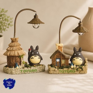 Könnte beinhalten: Zwei dekorative Lampen mit Totoro-Figuren. Die linke Lampe hat eine Hütte mit Strohdach und einen gebogenen Arm mit Licht. Die rechte Lampe zeigt eine Holzhütte mit Schornstein und ein ähnliches Licht. Beide Sockel sind steinfarben mit dem Text TOTORO.