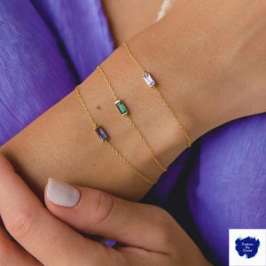 Può includere: Tre delicati braccialetti a catena dorata, ciascuno con una gemma rettangolare di diversi colori: viola, verde e viola chiaro. I braccialetti sono esposti su un polso su sfondo viola.