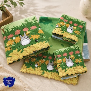 Può includere: Un set di asciugamani verdi con personaggi Totoro ricamati in grigio e bianco, piccoli spiritelli della fuliggine neri e motivi floreali gialli. Gli asciugamani presentano un bordo smerlato giallo e un logo blu Custom By Conor.