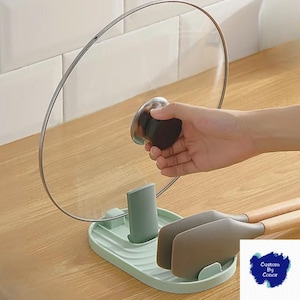 May include: A light green kitchen utensil rest with a holder for a pot lid and two spatulas. The rest is on a wooden surface. The pot lid is clear with a silver rim.