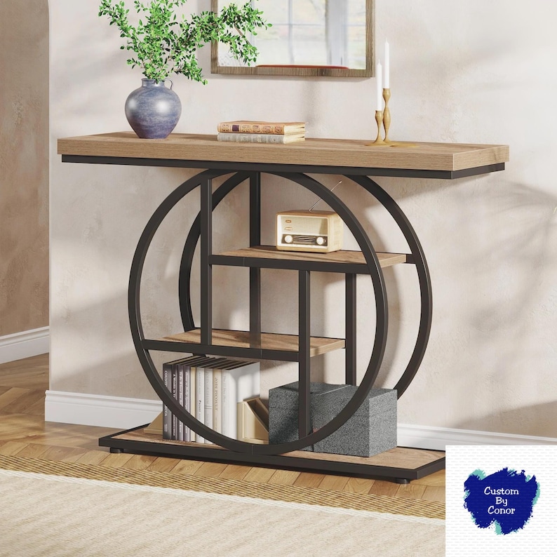 Peut inclure: Une console moderne avec un plateau et des &eacute;tag&egrave;res en bois brun clair, soutenue par une structure en m&eacute;tal noir avec des cercles d&eacute;coratifs. Elle pr&eacute;sente une petite radio vintage, des livres et des objets d&eacute;coratifs. Un vase bleu avec de la verdure est pos&eacute; dessus.