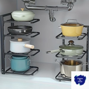 May include: A black metal pot and pan organiser with multiple shelves, holding various pots and pans in different colours. The pots include a light green pot, a black frying pan, a white pot with a wooden handle, and a teal pot. The organiser is designed to fit under a sink.