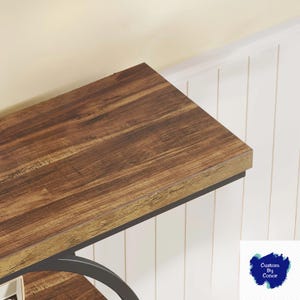 Peut inclure: Gros plan sur une table en bois avec une finition marron fonc&eacute;. La table a un cadre en m&eacute;tal noir et est plac&eacute;e contre un mur &agrave; panneaux blancs. La surface de la table pr&eacute;sente un motif de grain de bois.
