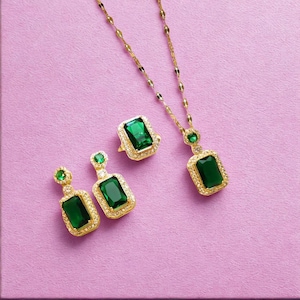 Op de afbeelding: Een goudkleurige sieradenset met een ketting, ring en oorbellen, elk versierd met een grote, rechthoekige smaragdgroene steen. De stenen zijn gezet in een gouden montuur, met kleinere accentstenen langs de randen. De achtergrond is effen roze.