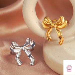 Peut inclure: Deux bagues en forme de nœud papillon, une argentée et une dorée, sont présentées sur un fond rose clair. Les bagues présentent un design détaillé en trois dimensions, avec les boucles et les extrémités de l'arc clairement définies.