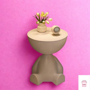 May include: A unique, beige-colored side table with a round top, shaped like a person. The table holds a small vase with yellow flowers and a small, round speaker. The background is a vibrant pink.