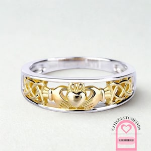 Puede incluir: Un anillo de Claddagh con una banda plateada y un diseño dorado de corazón, manos y corona. El anillo presenta un diseño calado con detalles de nudos celtas. Símbolo de amor, lealtad y amistad.