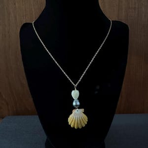 May include: A delicate necklace featuring a small, natural seashell pendant. The pendant is a light yellow color with a fan-like design. A dark gray pearl and a white, heart-shaped charm are also included. The necklace is displayed on a black mannequin.