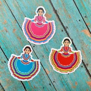 May include: Three colorful stickers featuring women in traditional Mexican dresses. Each sticker has a different color dress with a multi-colored ruffled skirt. The dresses are pink, red, and blue.
