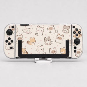 May include: A handheld gaming console featuring a cream-colored skin with a repeating pattern of small, cartoon-style cats in shades of brown, gray, and white, accented by tiny pink hearts, held in a black plastic stand against a plain background.