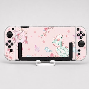 May include: A handheld gaming console featuring a light pink skin with illustrations of two dragons, cherry blossom branches, and a butterfly. The console has black buttons and joysticks on both sides, and it is mounted on a black and white stand.