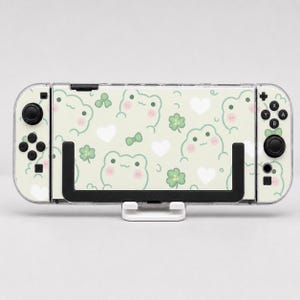 May include: A handheld gaming console featuring a light green skin with a repeating pattern of small smiling frogs, white hearts, green four-leaf clovers, and small bows. The device has black joysticks and buttons, and it rests on a white stand.