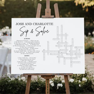 May include: A white sign on a wooden easel with the text "Josh and Charlotte Sip & Solve" and a crossword puzzle. The sign is outdoors, with a blurred background of a wedding reception.