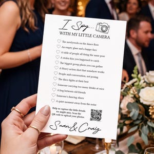 May include: A white rectangular wedding I Spy game card with black text and a camera icon. The card lists photo prompts, a QR code for photo uploads, and the names Swan & Craig. It is held against a background of a wedding reception dinner table.