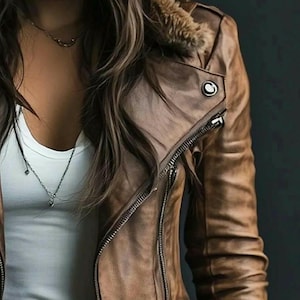 Puede incluir: Chaqueta de cuero marr&oacute;n con cuello de piel sint&eacute;tica y cremallera diagonal. La chaqueta tiene un bot&oacute;n en el hombro y una cremallera plateada. La modelo lleva una camiseta sin mangas blanca y un collar plateado.